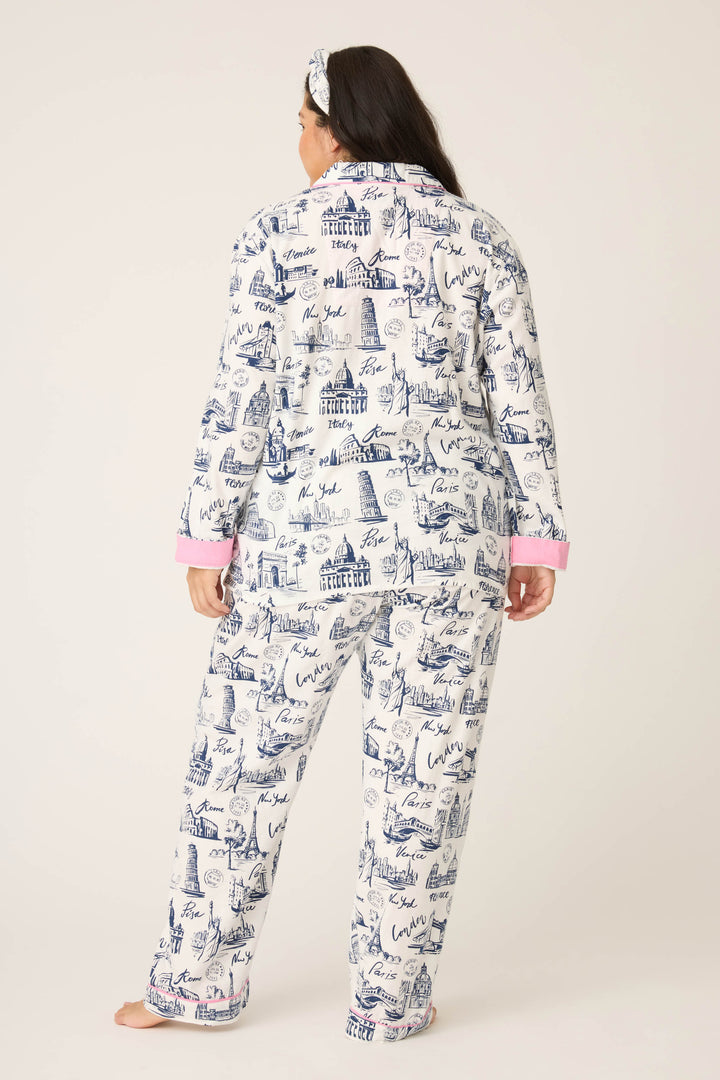 A person stands barefoot against a plain, light background wearing P.J. Salvages 3-Piece Set WILD PJ SET FLANNELS PLUS: white pajamas with a blue cityscape print, pink cuffs, long sleeves and pants, plus a matching headband.