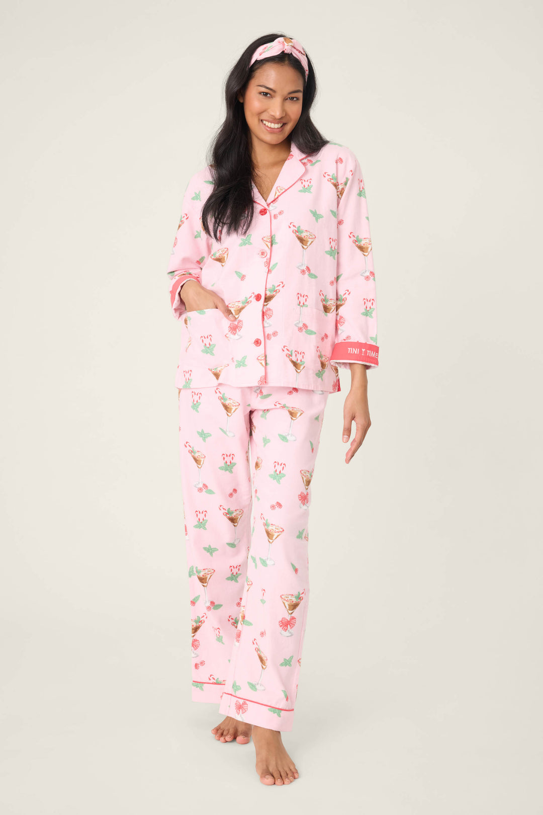 A woman stands barefoot wearing the P.J. Salvage Tini Time Drink Print Flannel PJ Set, featuring matching pants and top with playful drink prints, plus a coordinating headband and pink piping accents.