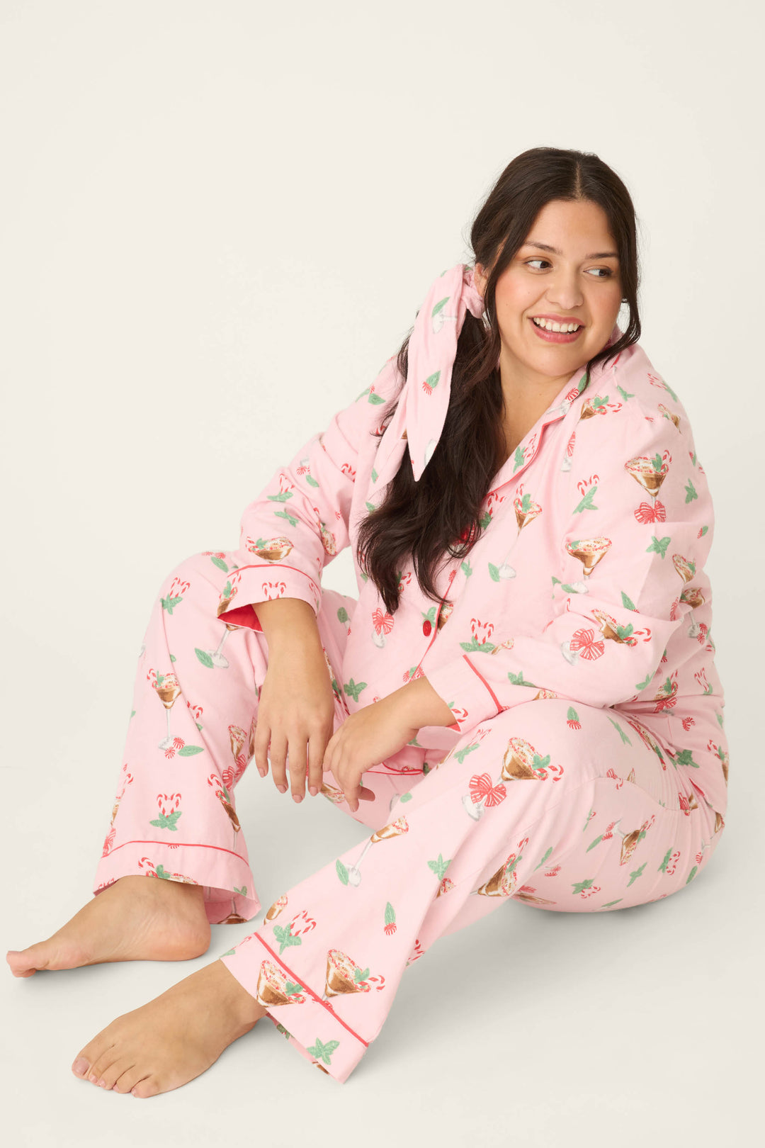 A person with long dark hair sits on the floor, smiling, in the P.J. Salvage Tini Time Drink Print Flannel Plus Size PJ Set with a matching pink hair bow. The background is plain and light-colored.