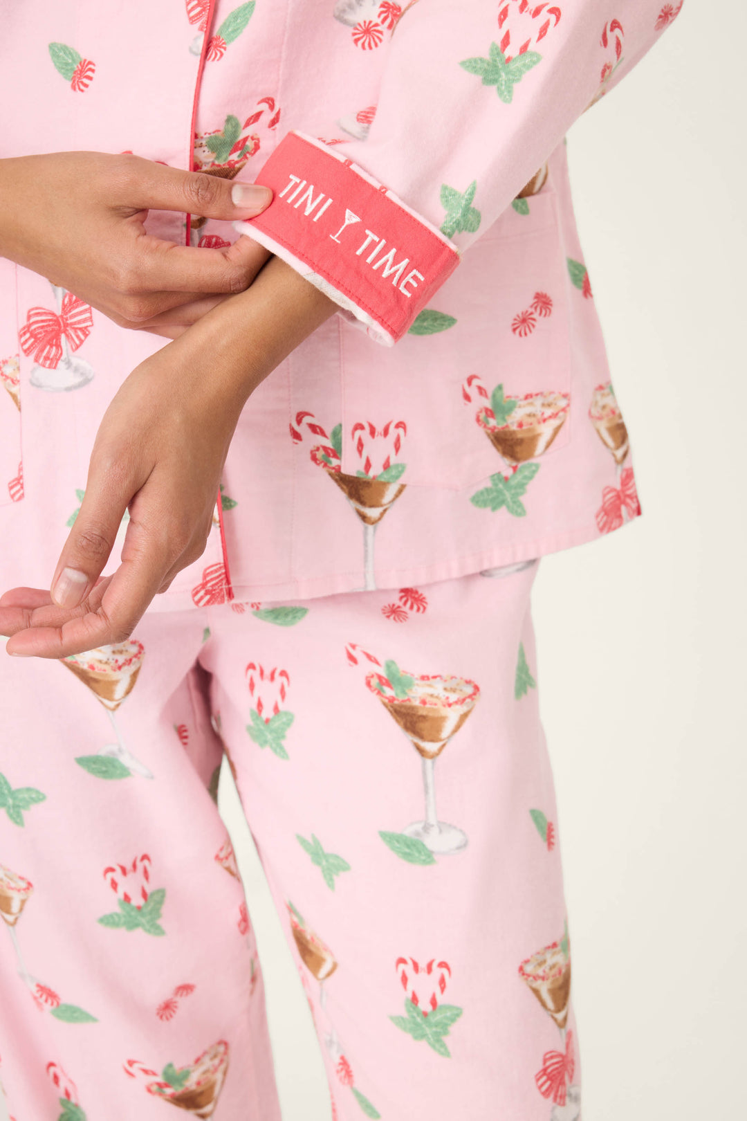 A person in the P.J. Salvage Tini Time Drink Print Flannel PJ Set adjusts their sleeve, revealing embroidered "TIN I TIME" text on pink pajamas decorated with candy canes and cocktail glasses.