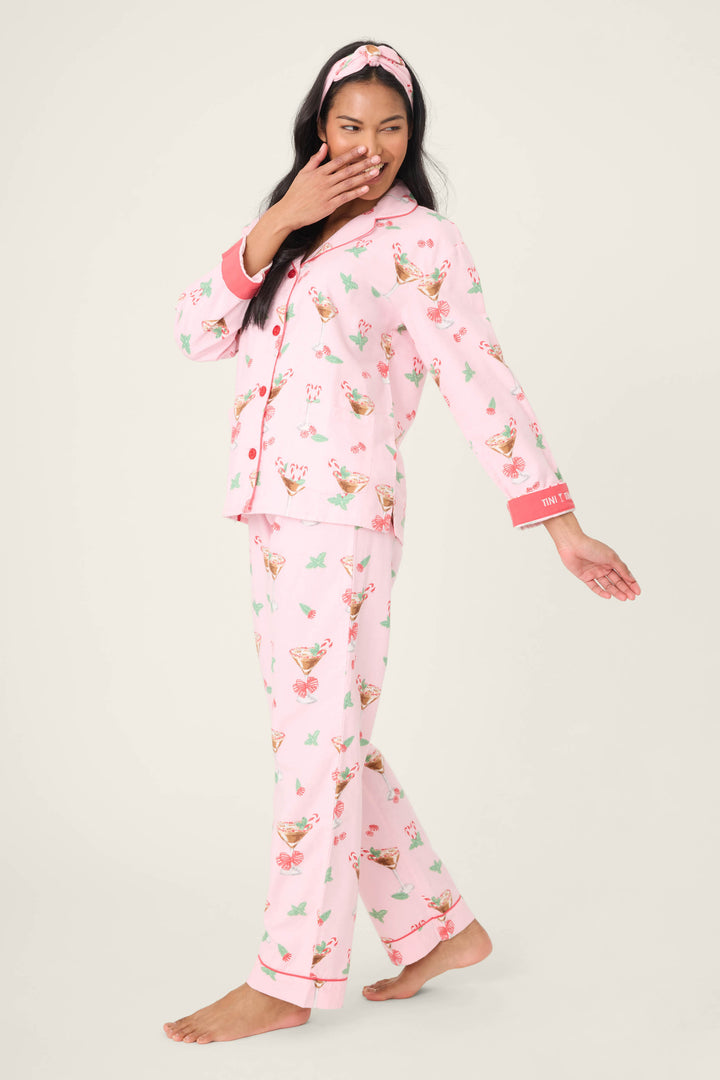 A woman smiles barefoot on a light background, wearing the P.J. Salvage Tini Time Drink Print Flannel PJ Set and matching headband, with one hand near her mouth and the other arm extended.