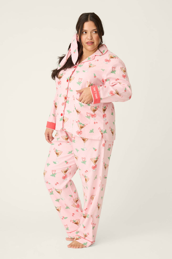 A woman with long dark hair poses barefoot in the P.J. Salvage Tini Time Drink Print Flannel Plus Size PJ Set, paired with a matching bow, standing against a plain off-white background.