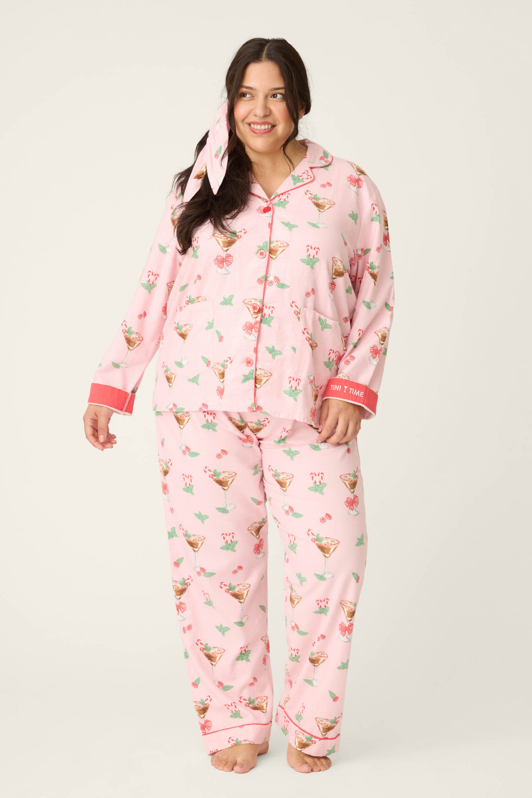 A woman with long dark hair models the Tini Time Drink Print Flannel Plus Size PJ Set by P.J. Salvage, featuring light pink pajamas with cocktail glass print, collared button-up top, loose-fitting pants, and matching headband against a plain background.