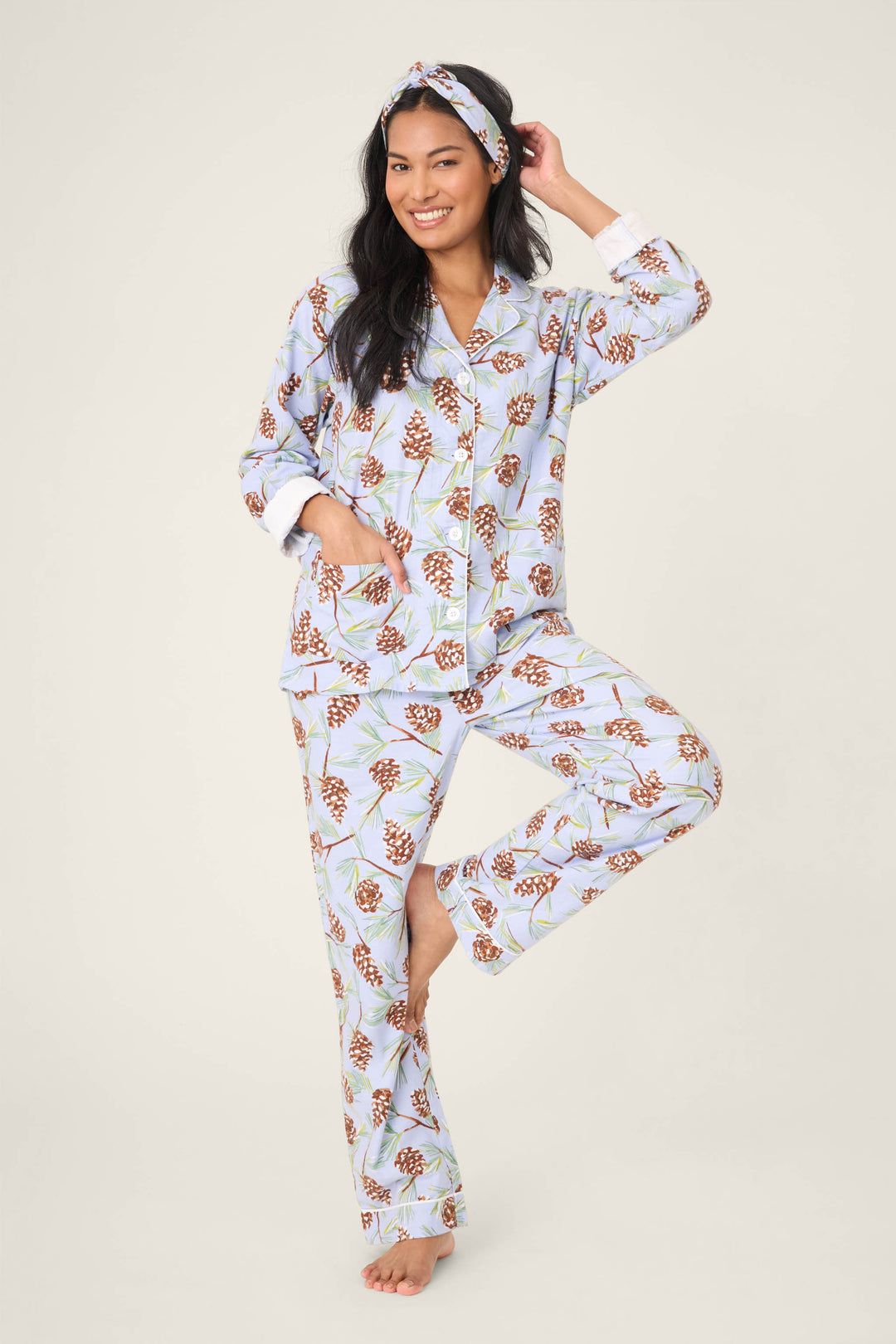 A woman balances on one leg, smiling, in the P.J. Salvage Feelin' Pine Pinecone Print PJ Set—featuring a matching light blue pajama set and headband with brown pinecones and green leaves, against a plain backdrop.