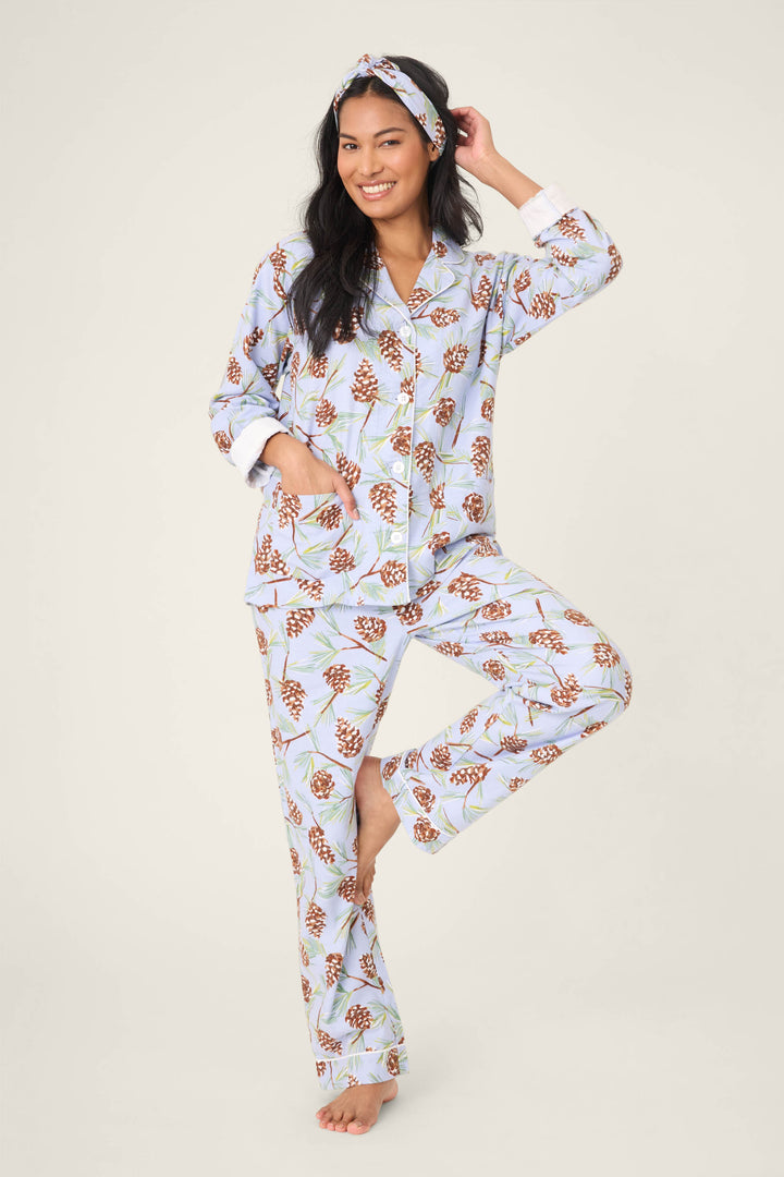 A woman balances on one leg, smiling, in the P.J. Salvage Feelin' Pine Pinecone Print PJ Set—featuring a matching light blue pajama set and headband with brown pinecones and green leaves, against a plain backdrop.