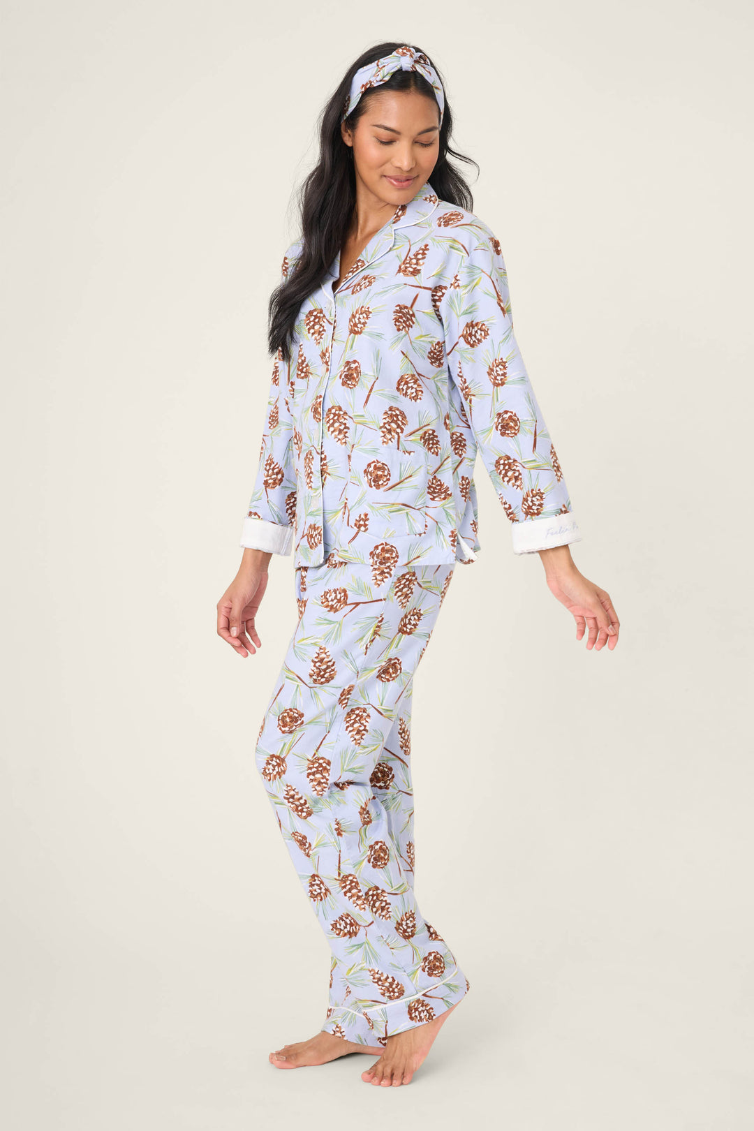 A woman models the Feelin' Pine Pinecone Print PJ Set by P.J. Salvage, featuring light blue pajamas with a brown pinecone pattern and a matching headband, standing barefoot against a plain light background.