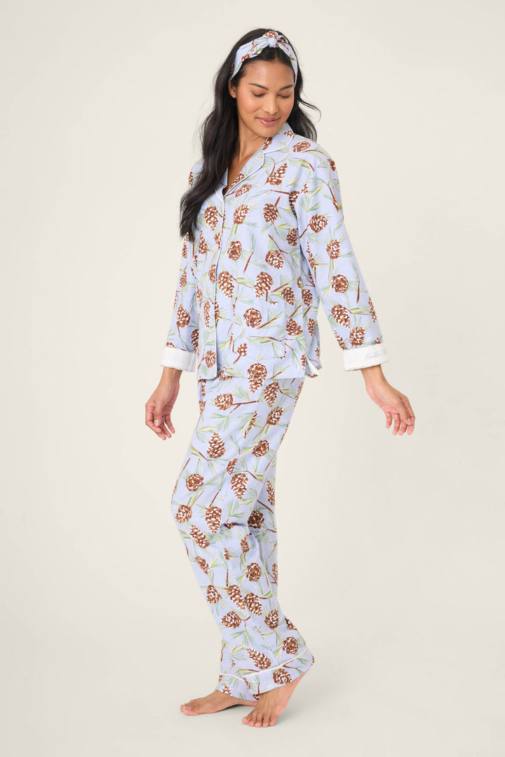 A woman models the Feelin' Pine Pinecone Print PJ Set by P.J. Salvage, featuring light blue pajamas with a brown pinecone pattern and a matching headband, standing barefoot against a plain light background.