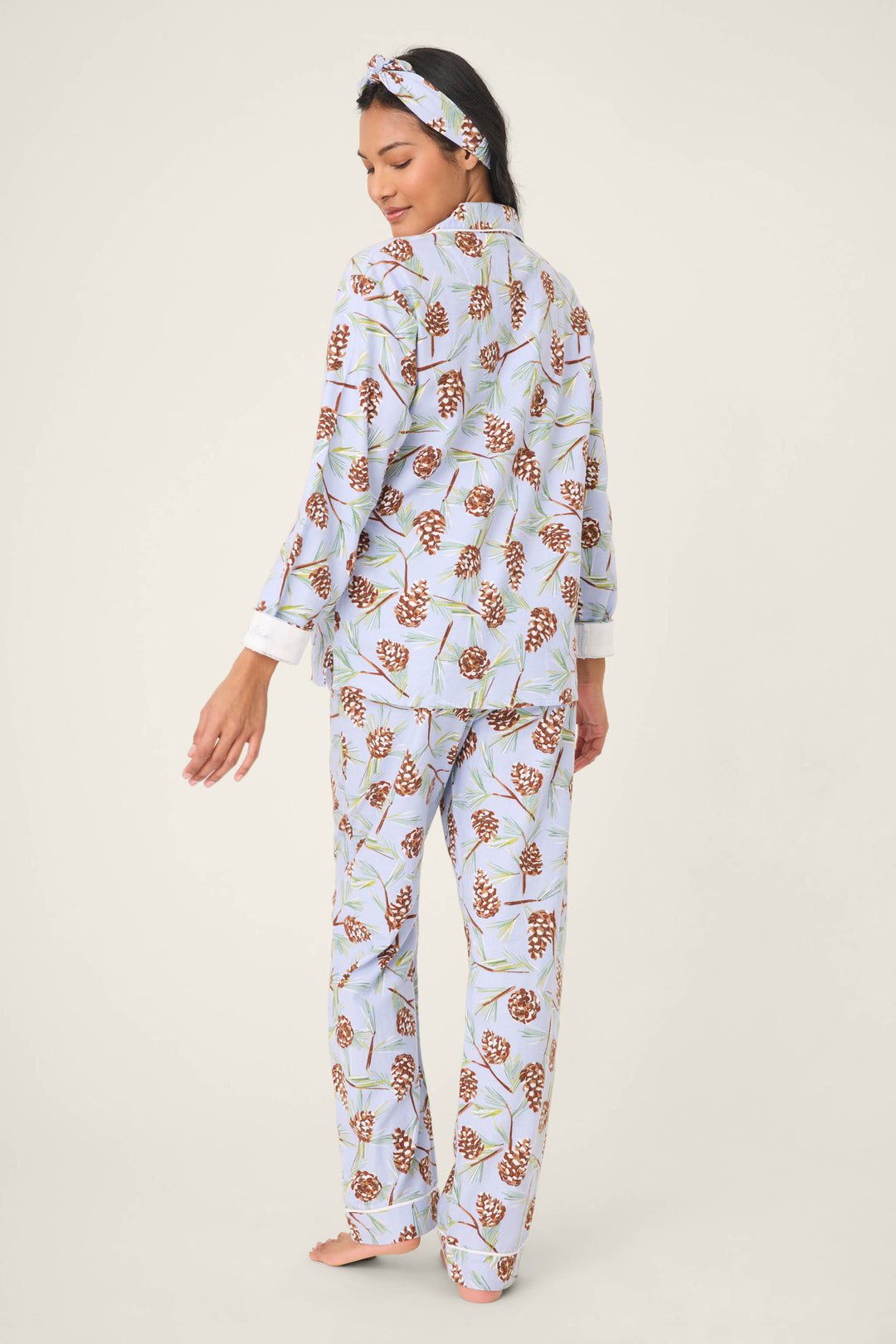 A woman faces away, barefoot, in the P.J. Salvage Feelin' Pine Pinecone Print PJ Set—a matching long-sleeve top and full-length pants with a botanical pine cone design on a light background.