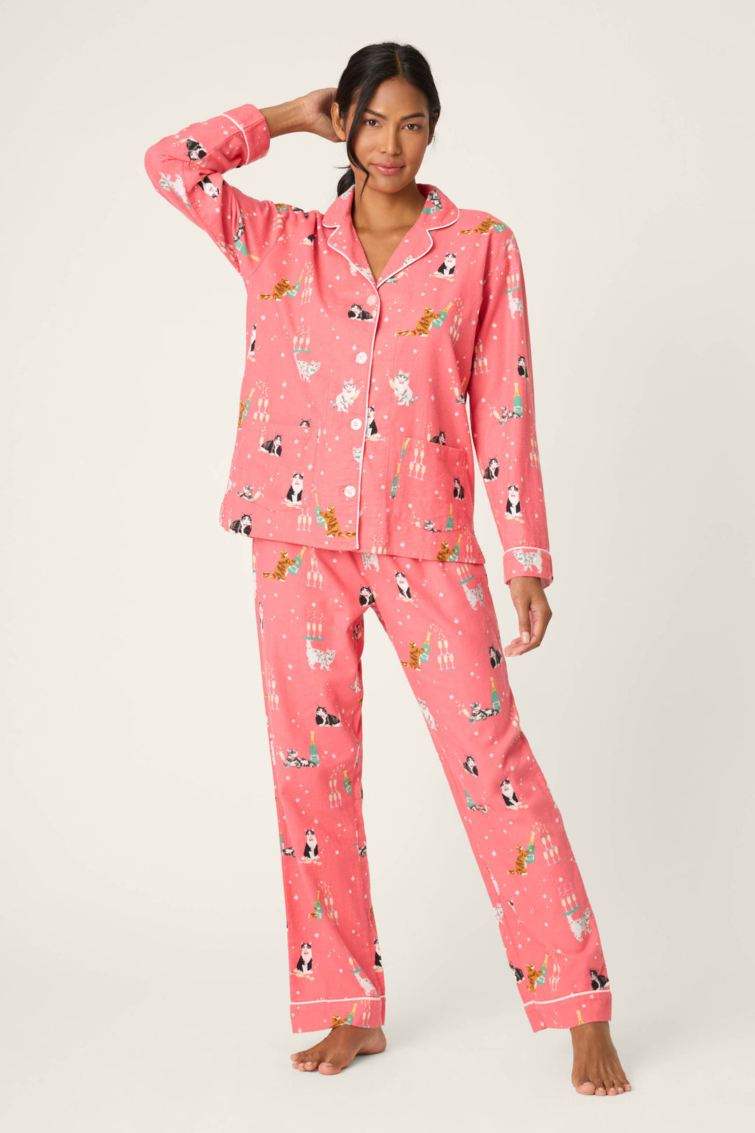A woman poses against a plain background in the P.J. Salvage Let's Get Fizzical Cat Print Flannel PJ Set, featuring a festive pink cat-print long-sleeve button-up top and matching pants, with one hand raised to her hair.