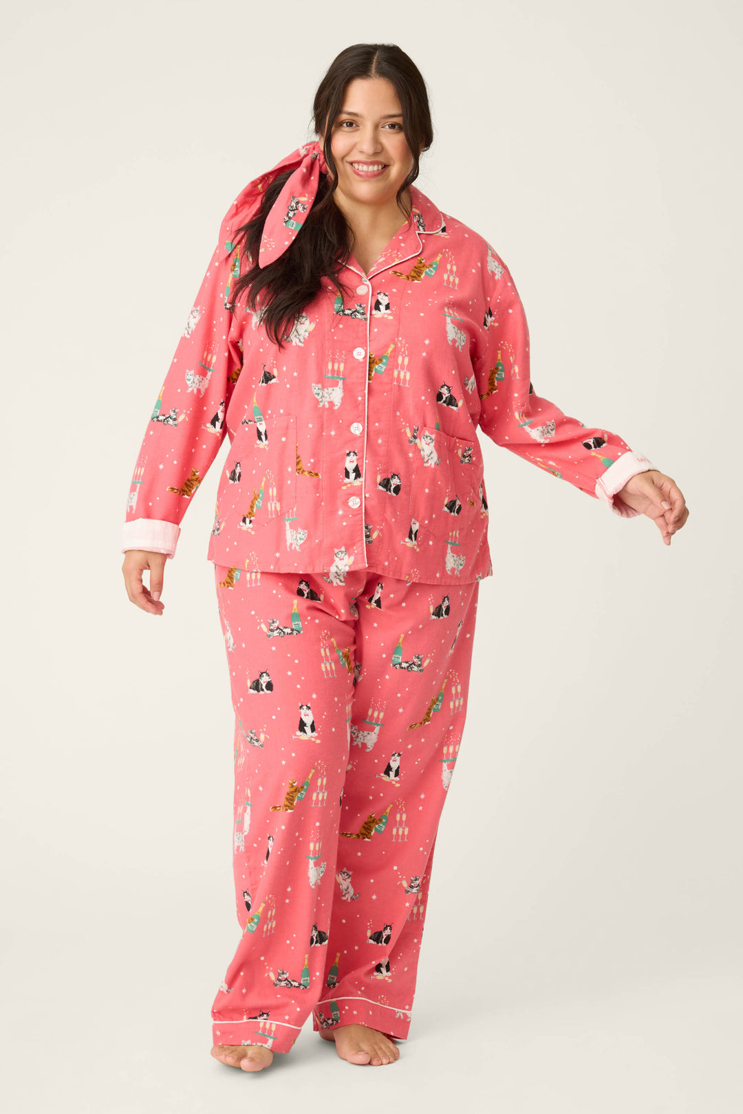A woman smiles barefoot on a light background, wearing the P.J. Salvage Let's Get Fizzical Cat Print Flannel Plus Size PJ Set with white trim and a matching pink hair tie, one hand slightly raised.