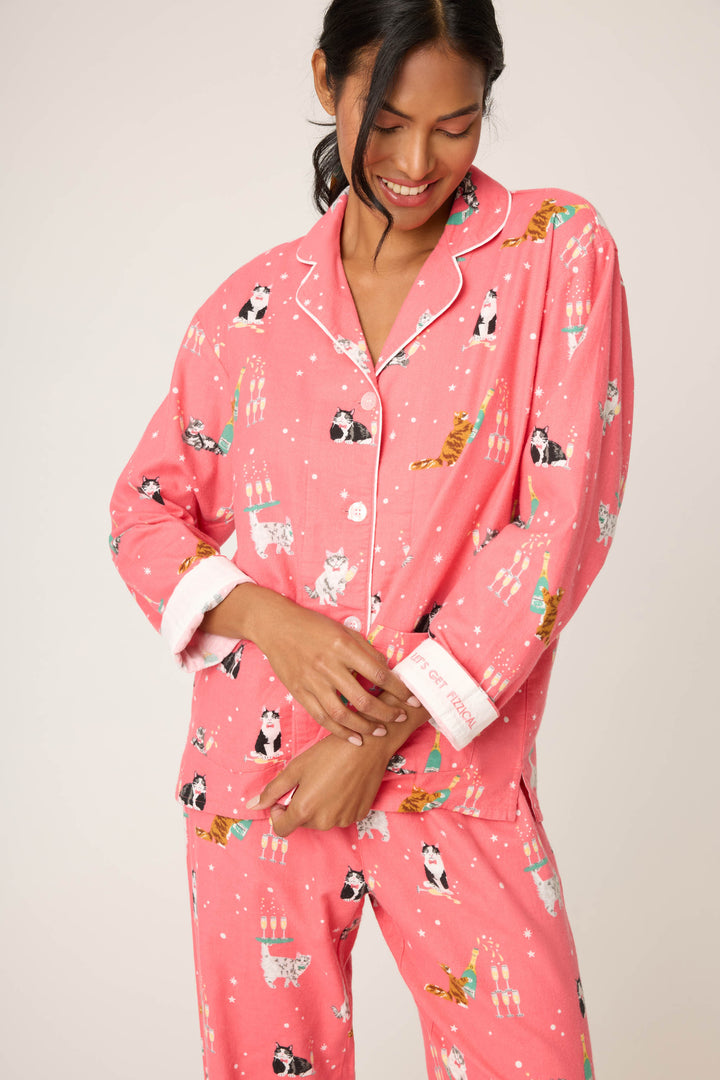 A person wears the P.J. Salvage Let's Get Fizzical Cat Print Flannel PJ Set, featuring playful cat and candle designs, standing against a plain background with a smile, head tilted down, and hands resting on the pajama top.