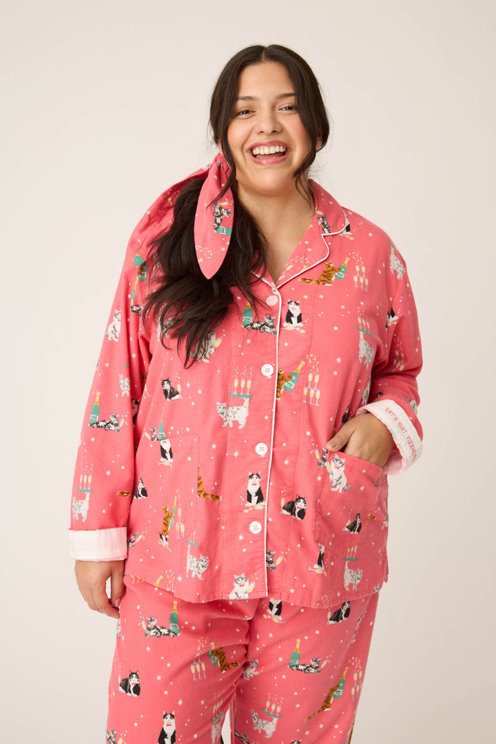 A woman with long dark hair smiles at the camera in the P.J. Salvage "Let's Get Fizzical Cat Print Flannel Plus Size PJ Set," featuring a cat and Christmas tree print and a matching bow, against a plain light background.