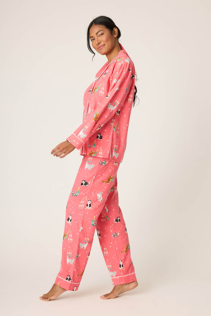 A person stands barefoot against a plain background, wearing the P.J. Salvage Let’s Get Fizzical Cat Print Flannel PJ Set—matching pink button-up long-sleeve shirt and pants decorated with cat and holiday-themed patterns.