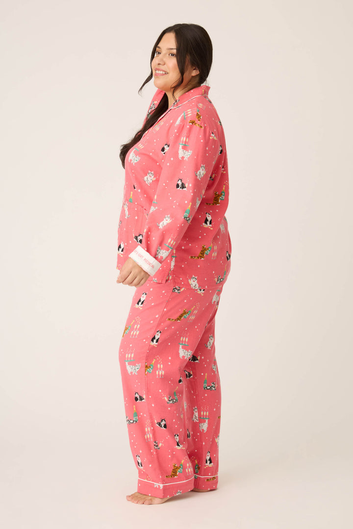 A woman with long dark hair stands barefoot, smiling slightly, in P.J. Salvage's Let's Get Fizzical Cat Print Flannel Plus Size PJ Set. She faces sideways against a plain off-white background.