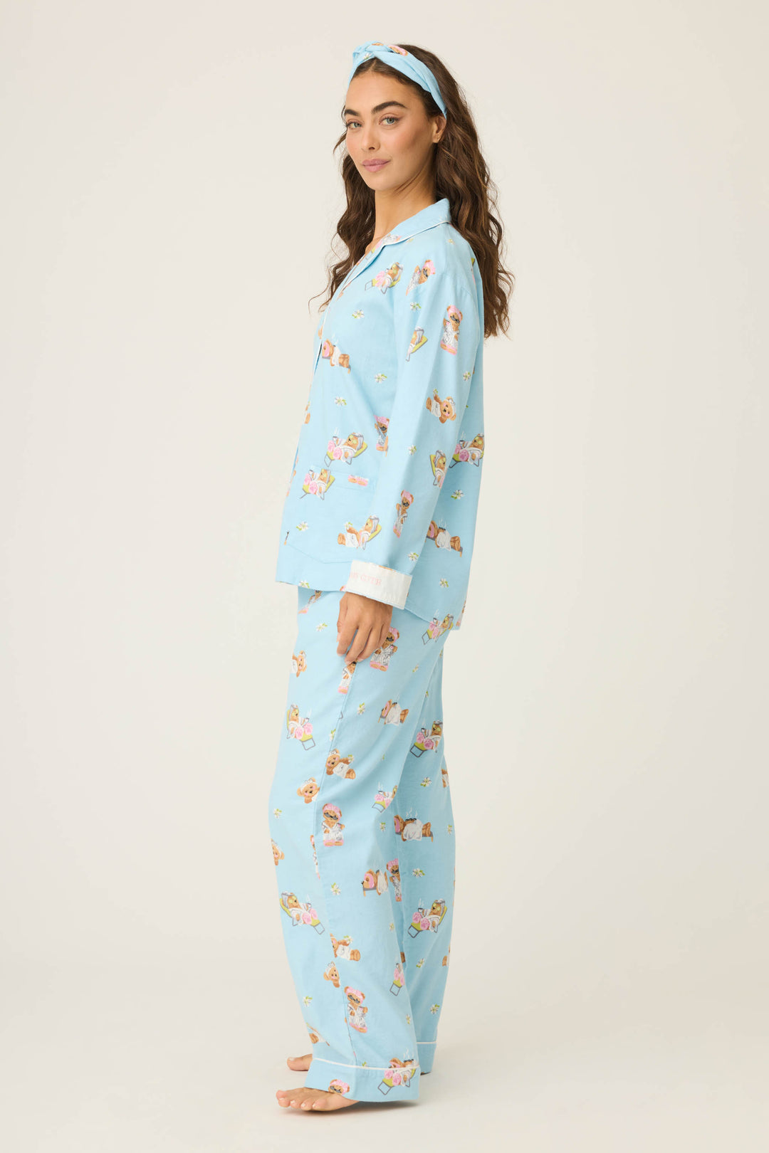 A person stands barefoot in profile, wearing the P.J. Salvage Beary Cute Bear Print Flannel PJ Set—light blue pajamas with a playful bear print, featuring a long-sleeve button-up top and loose pants—against a plain, light-colored background.