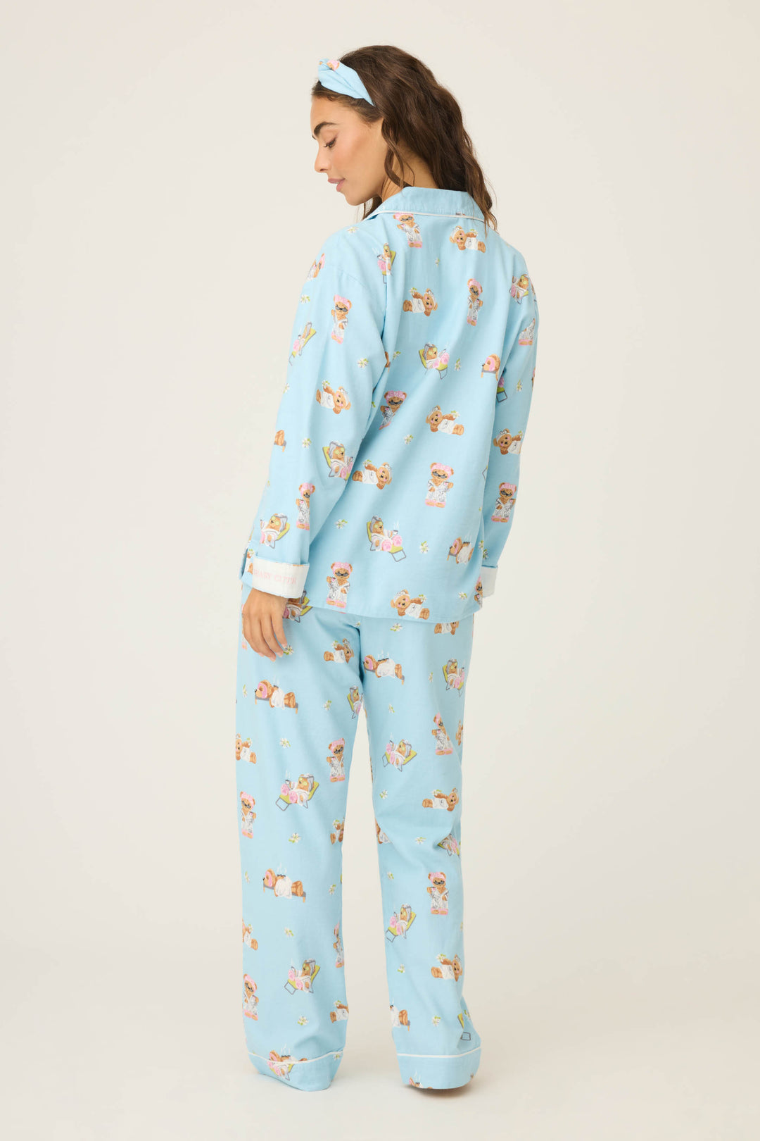 A woman faces away, wearing the P.J. Salvage Beary Cute Bear Print Flannel PJ Set—light blue, cartoon bear print, long-sleeve button-up top, loose pants, and matching headband—against a plain light background.