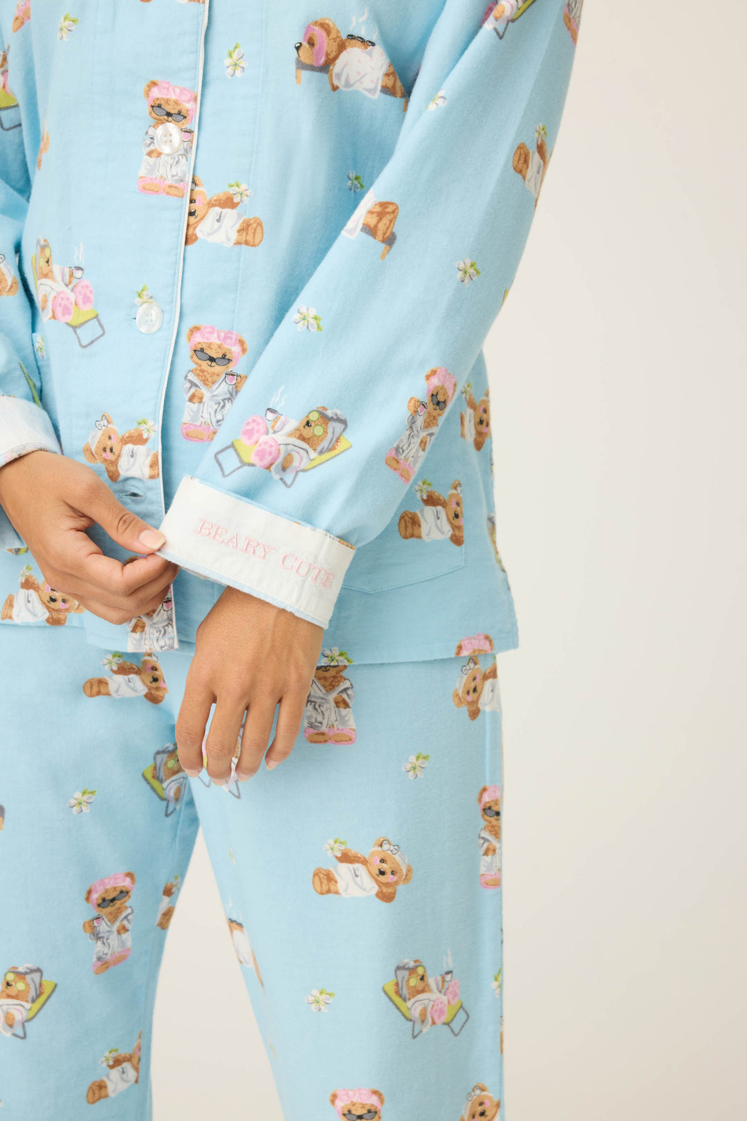 A person wears the P.J. Salvage Beary Cute Bear Print Flannel PJ Set in light blue, featuring bear and spa-themed patterns. One hand holds a sleeve cuff embroidered with BEARY CUTE in pink.