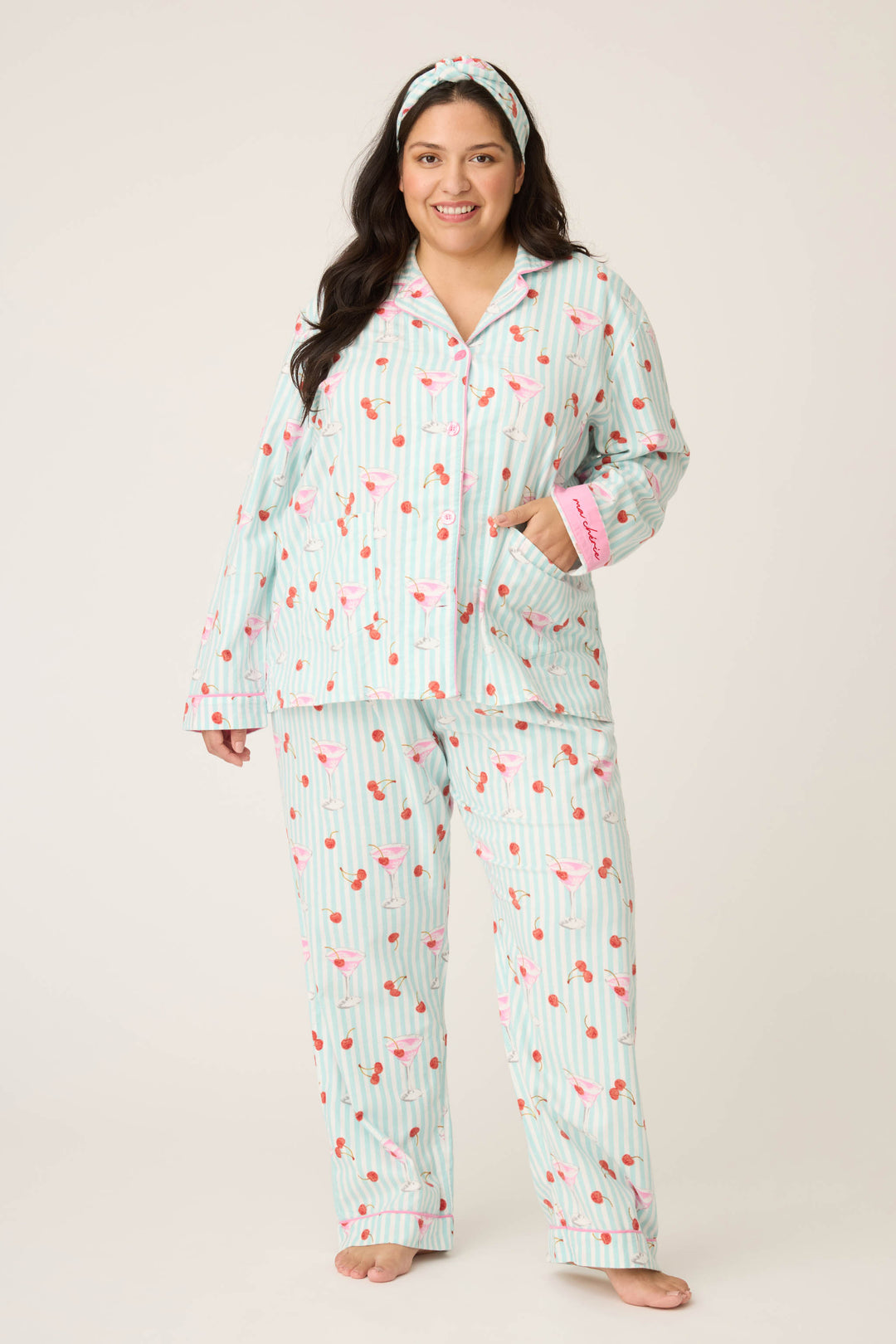 A woman with long dark hair and a matching headband smiles barefoot, wearing the PAISLEY PJ SET FLANNELS (Plus) by P.J. Salvage—a light blue and white striped cotton pajama set with a pink flamingo pattern.