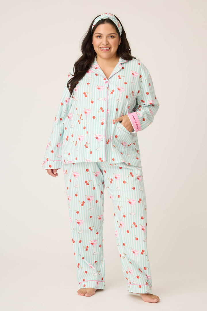 A woman with long dark hair and a matching headband smiles barefoot, wearing the PAISLEY PJ SET FLANNELS (Plus) by P.J. Salvage—a light blue and white striped cotton pajama set with a pink flamingo pattern.