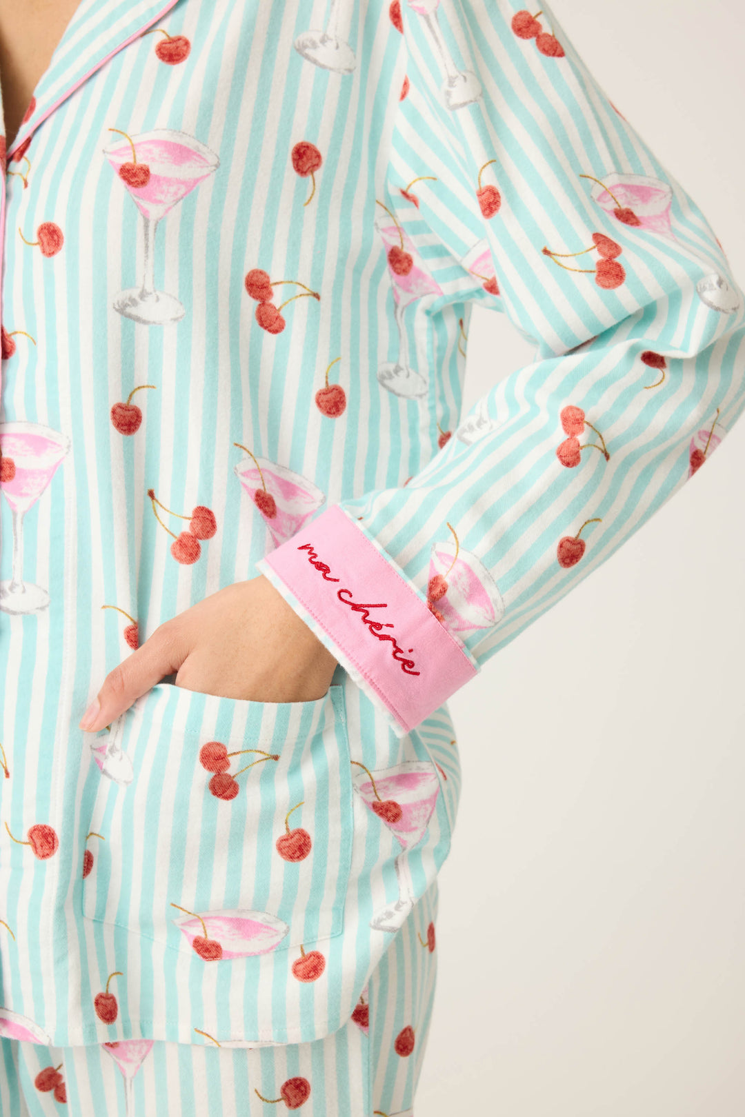 A person wears the P.J. Salvage 3-Piece Set PAISLEY PJ SET FLANNELS, featuring blue and white striped pajamas with cherry and cocktail glass prints, pink sleeve cuffs embroidered with “mon chéri,” and a hand in the pants pocket.