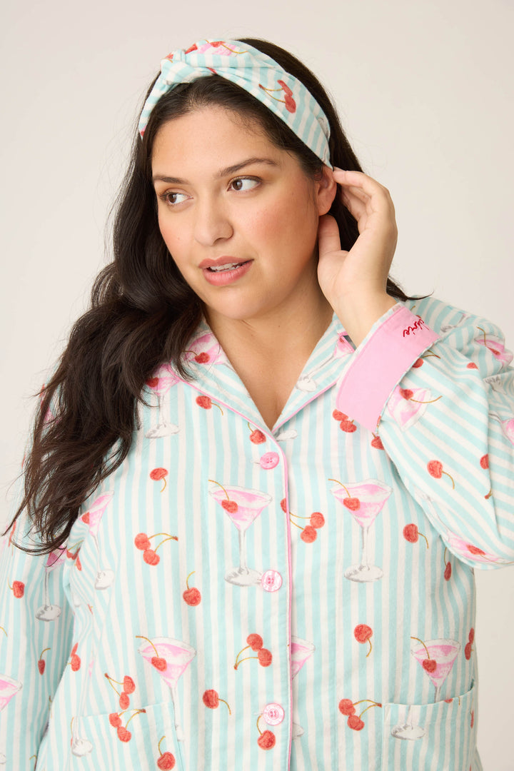 A woman wearing the PAISLEY PJ SET FLANNELS (Plus) from P.J. Salvage, featuring a light blue and white striped sleepwear top with pink martini and cherry prints and a matching headband, touches her ear while looking to the side.