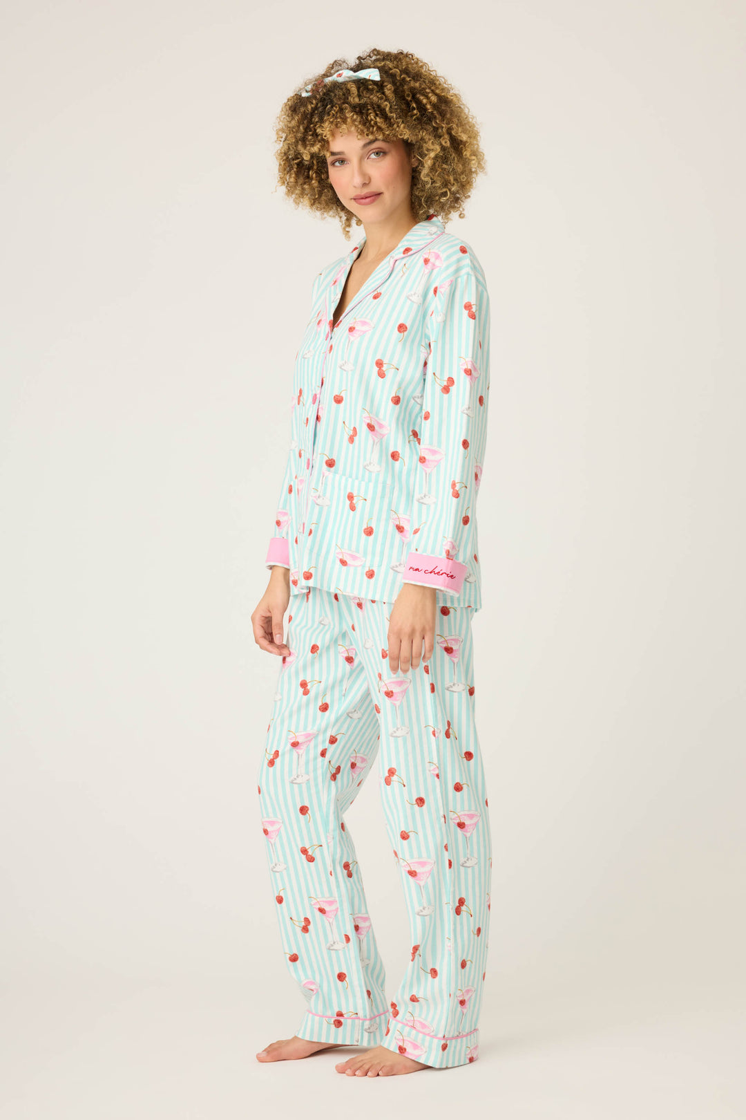 A person stands barefoot in P.J. Salvages 3-Piece Set PAISLEY PJ SET FLANNELS, featuring a light blue and white striped button-up top and long pants with cherry print and pink cuffs, against a plain light background.