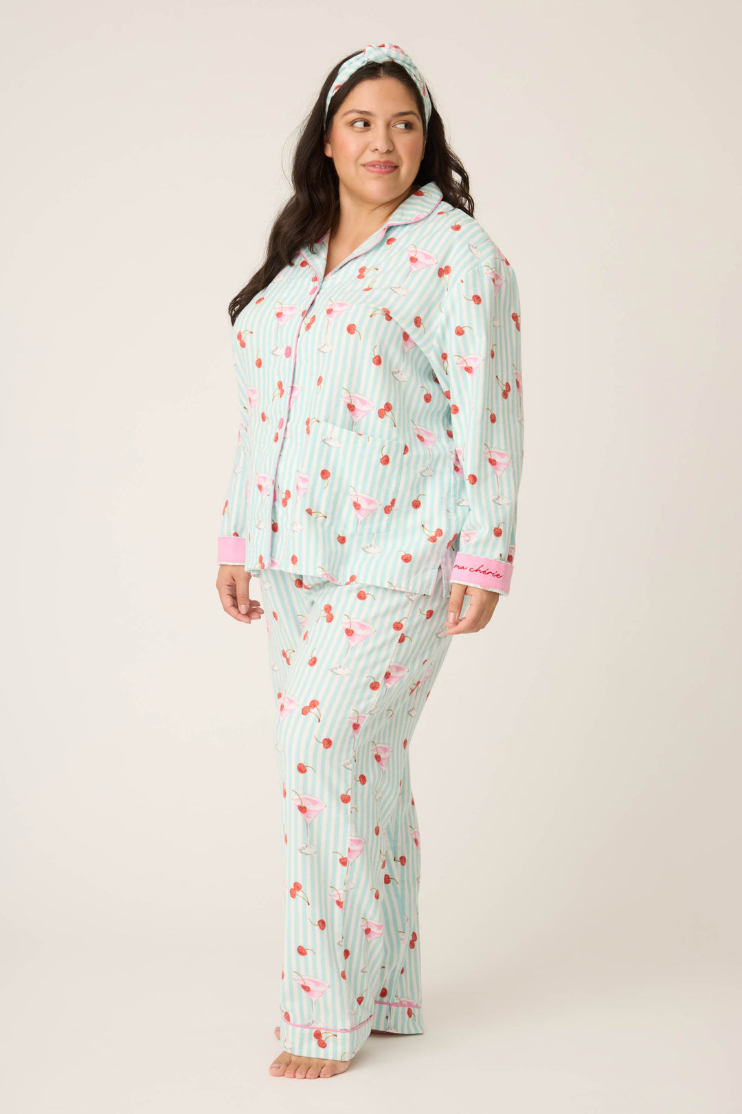 A woman in the PAISLEY PJ SET FLANNELS (Plus) by P.J. Salvage stands barefoot on a light background, wearing matching striped pajamas with flamingo and heart prints, a headband, and softly smiling as she looks to the side.