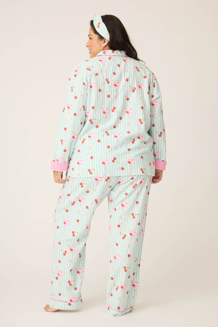 A woman wears the P.J. Salvage PAISLEY PJ SET FLANNELS (Plus)—a cozy flannel set with light blue stripes, pink flamingos, and heart patterns, plus pink cuffs and a matching headband—standing barefoot facing away against a plain background.