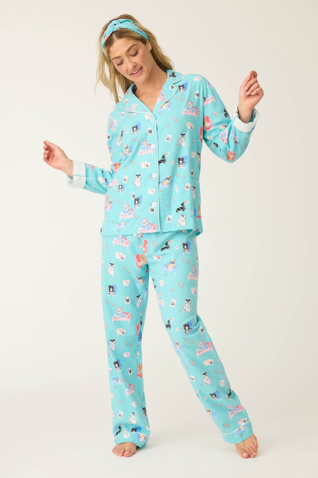 A person wears the P.J. Salvage This Is How I Roll Dog Print Flannel PJ Set: light blue, long-sleeved button-up pajamas with a colorful dog print and matching headband, barefoot against a plain background.