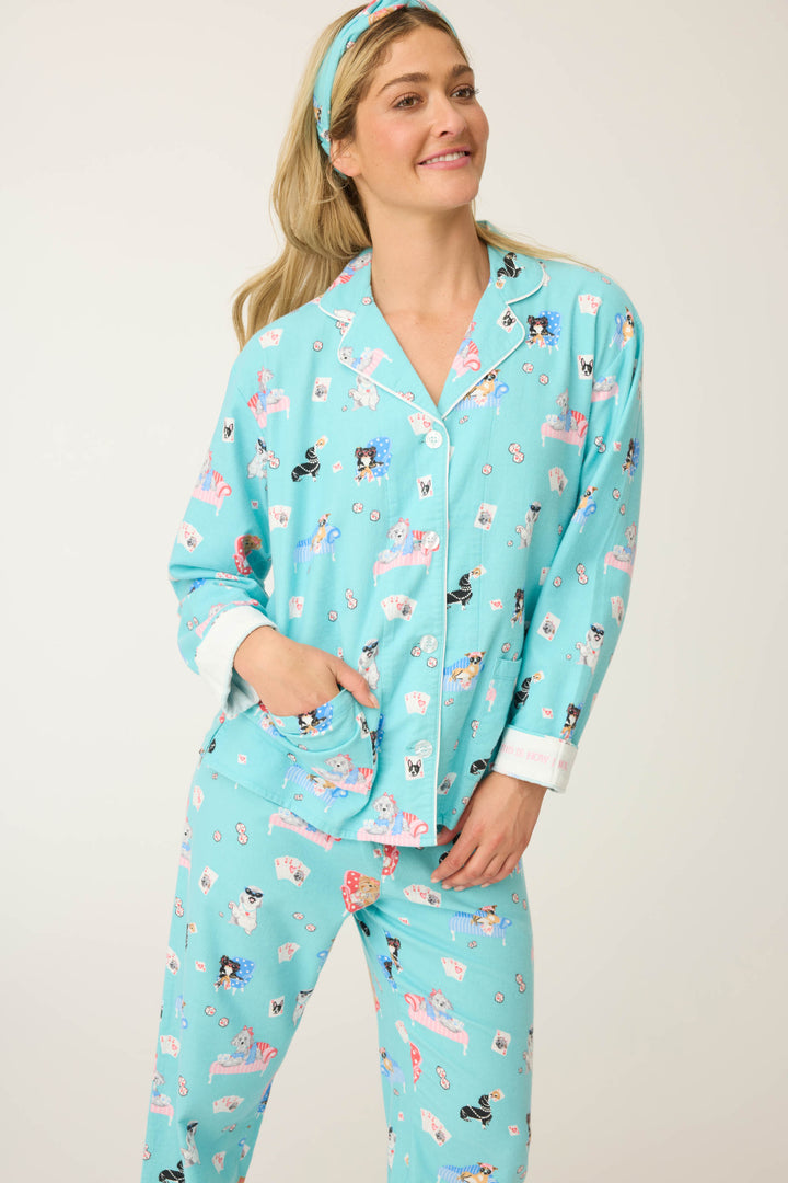 A person smiles in the P.J. Salvage This Is How I Roll Dog Print Flannel PJ Set, featuring matching light blue pajamas with a playful dog print and a coordinating headband, with one hand in their pocket against a plain background.