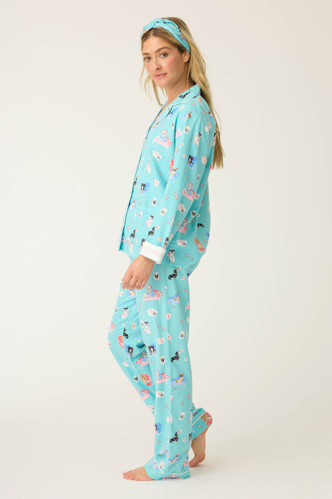 A woman stands barefoot in profile, wearing P.J. Salvages This Is How I Roll Dog Print Flannel PJ Set with a matching headband against a plain, light background.