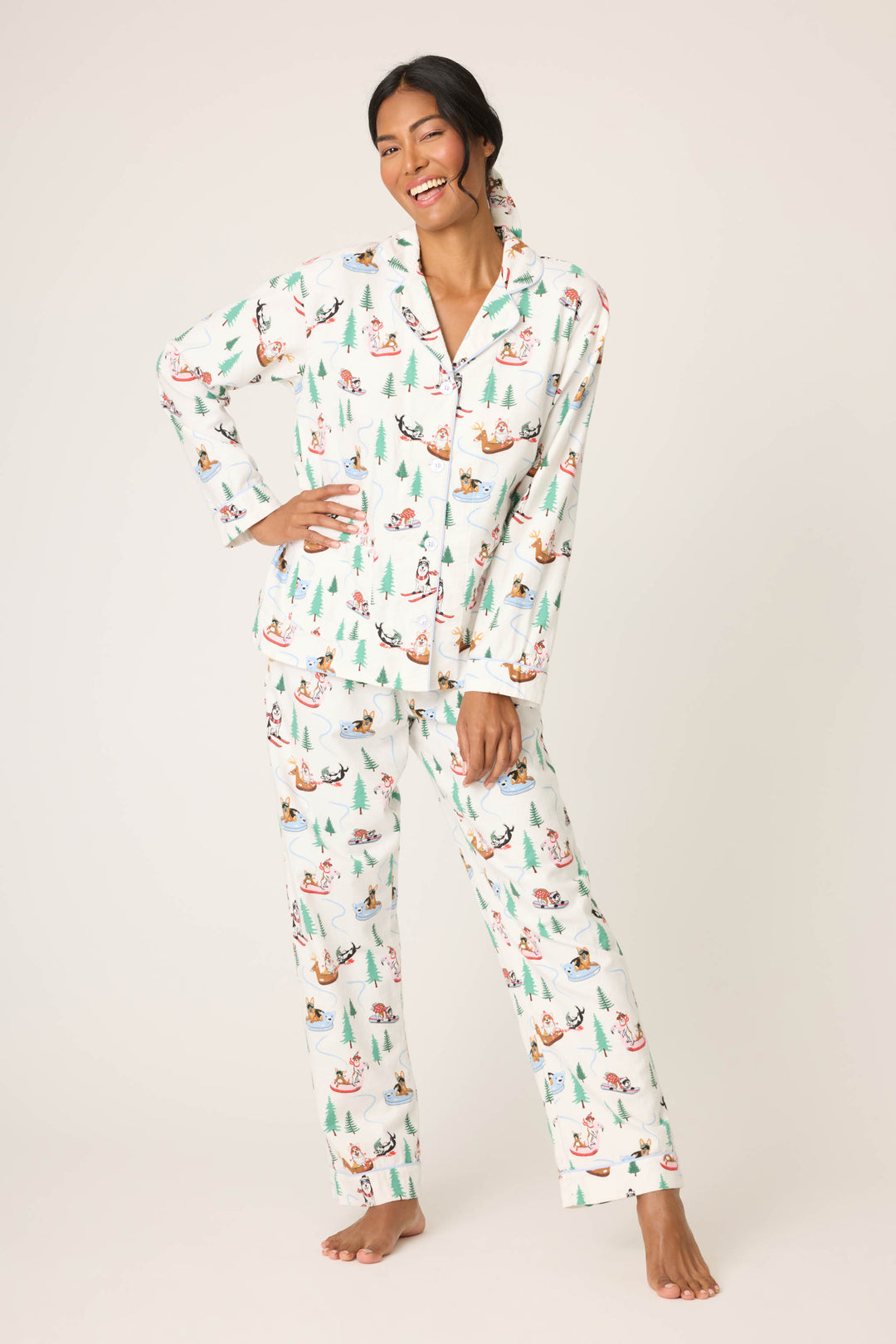 A woman, barefoot and smiling, models the P.J. Salvage Sleigh All Day Winter Print Flannel PJ Set with long sleeves and festive penguin and tree motifs, standing against a plain background with one hand touching her leg.