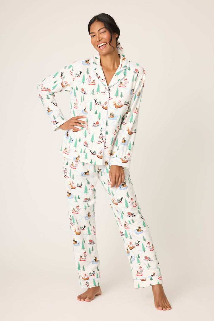 A woman, barefoot and smiling, models the P.J. Salvage Sleigh All Day Winter Print Flannel PJ Set with long sleeves and festive penguin and tree motifs, standing against a plain background with one hand touching her leg.