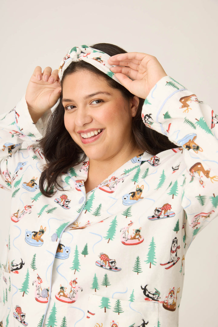 A woman smiles at the camera while adjusting her headband, wearing the P.J. Salvage Sleigh All Day Winter Print Flannel Plus Size PJ Set with a matching headband against a plain, light background.