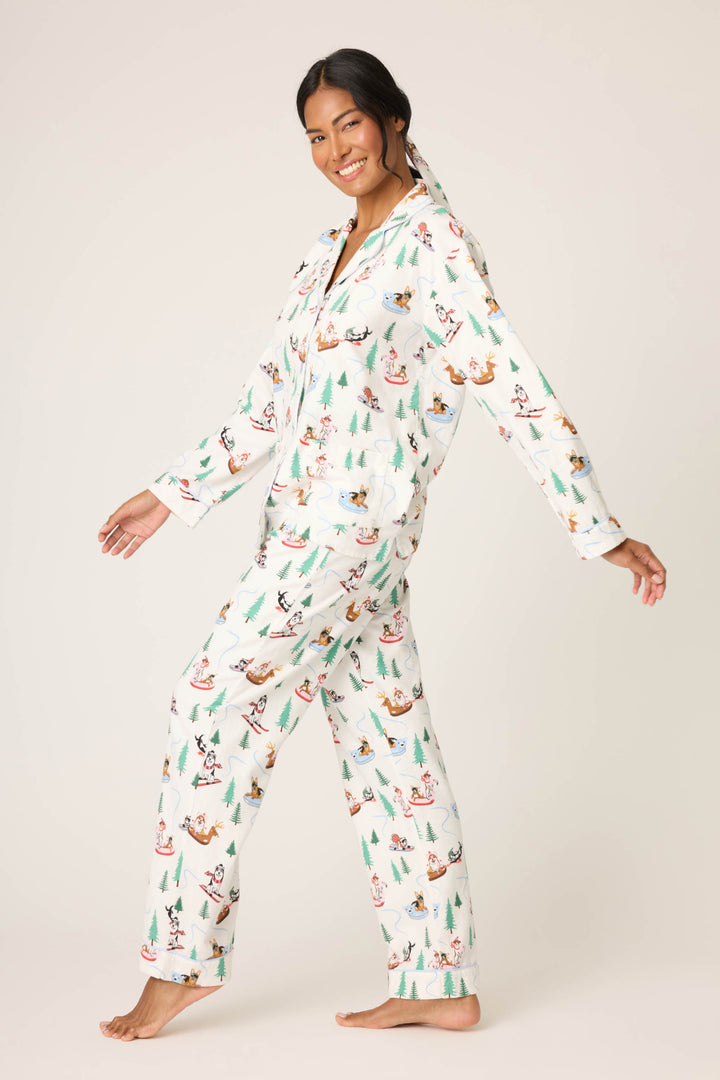 A person stands barefoot against a light background, smiling and wearing the P.J. Salvage Sleigh All Day Winter Print Flannel PJ Set with festive trees and animals. They pose with one foot slightly lifted.