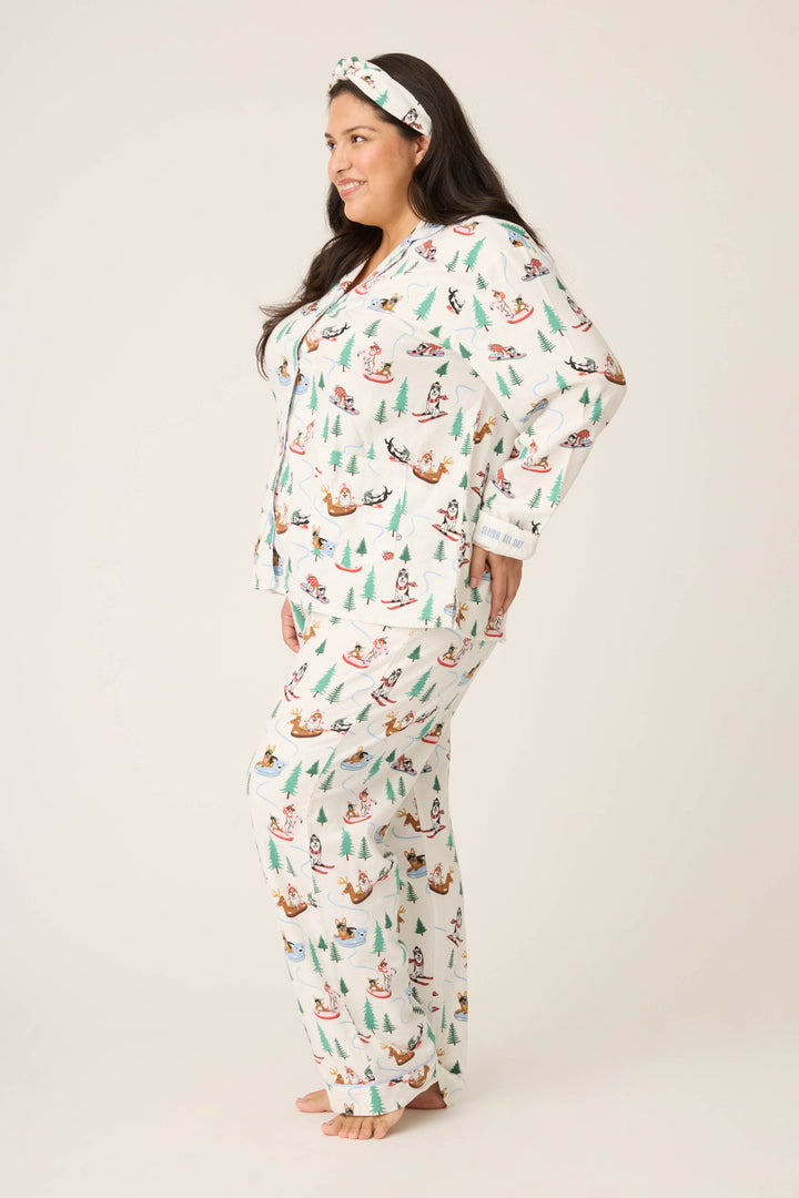 A woman stands barefoot in profile, wearing the P.J. Salvage "Sleigh All Day" Winter Print Flannel Plus Size PJ Set—white pajamas and headband featuring festive dogs, Christmas trees, and sleds—against a plain, light-colored background.