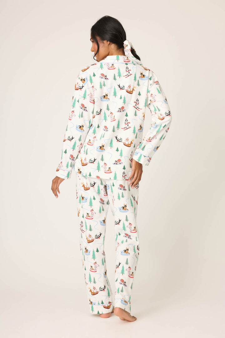 A woman stands barefoot facing away, wearing the Sleigh All Day Winter Print Flannel PJ Set by P.J. Salvage, featuring a colorful pattern of trees and dogs. Her hair is tied back with a matching scrunchie against a plain off-white background.