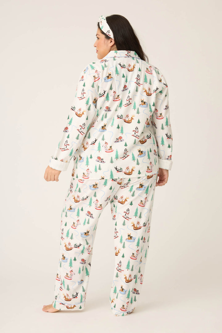 A person stands barefoot facing away, wearing the P.J. Salvage Sleigh All Day Winter Print Flannel Plus Size PJ Set, which features a festive pattern with trees, sleds, and animals. The background is plain and light-colored.