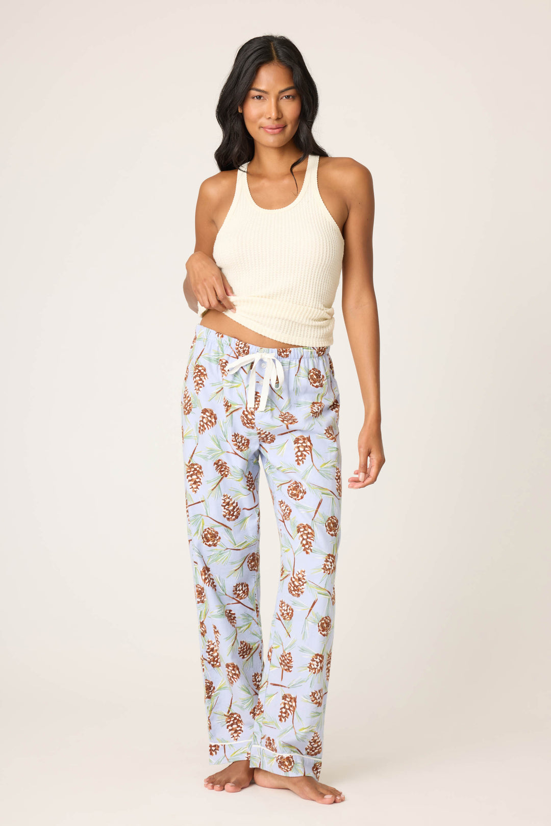 A woman with long dark hair stands barefoot against a plain background, wearing a cream ribbed tank top and P.J. Salvage Feelin' Pine Pinecone Print Flannel Pants with a drawstring waist. She smiles slightly.