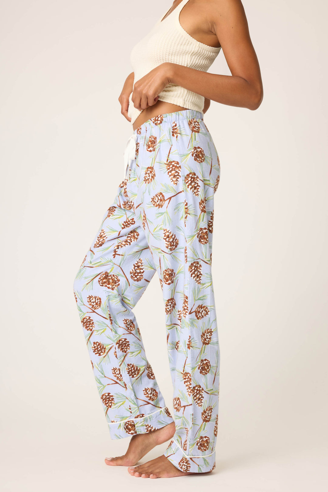 A person wearing a sleeveless, ribbed white tank top and P.J. Salvage Feelin' Pine Pinecone Print Flannel Pants stands barefoot against a plain background.