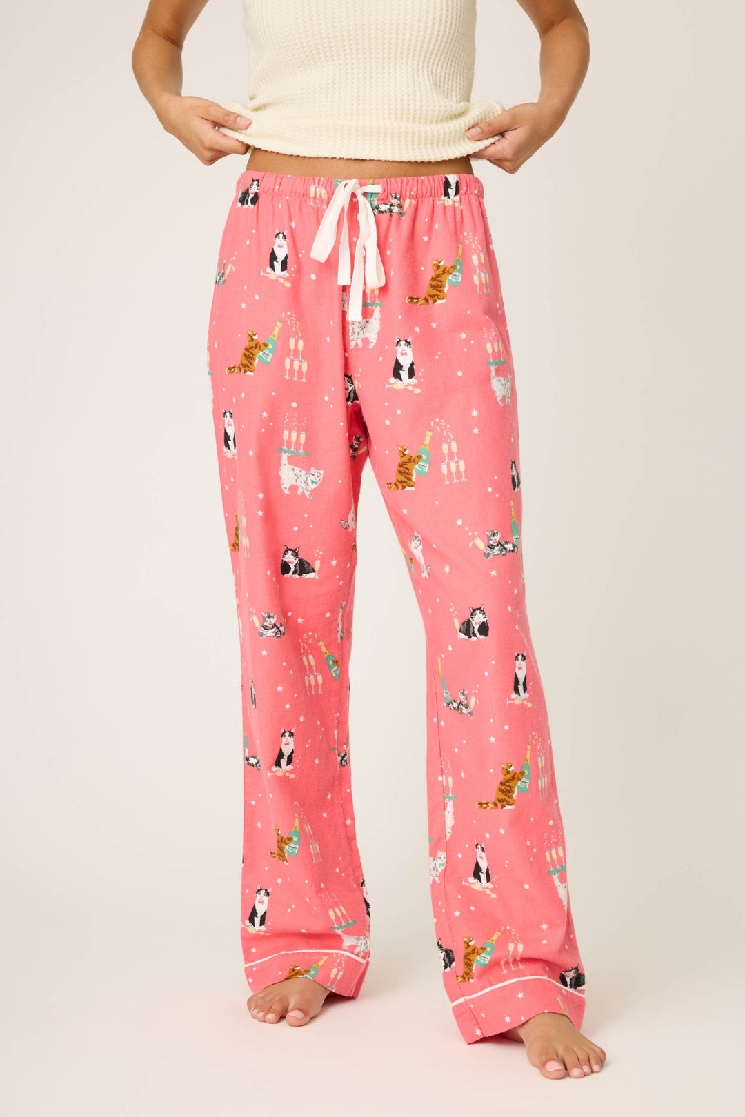A person adjusts the waistband of the P.J. Salvage Let’s Get Fizzical Cat Print Flannel Pant, featuring a pink color with cat, birthday cake, and candle pattern, white drawstring, and bare feet; the top is partially visible.