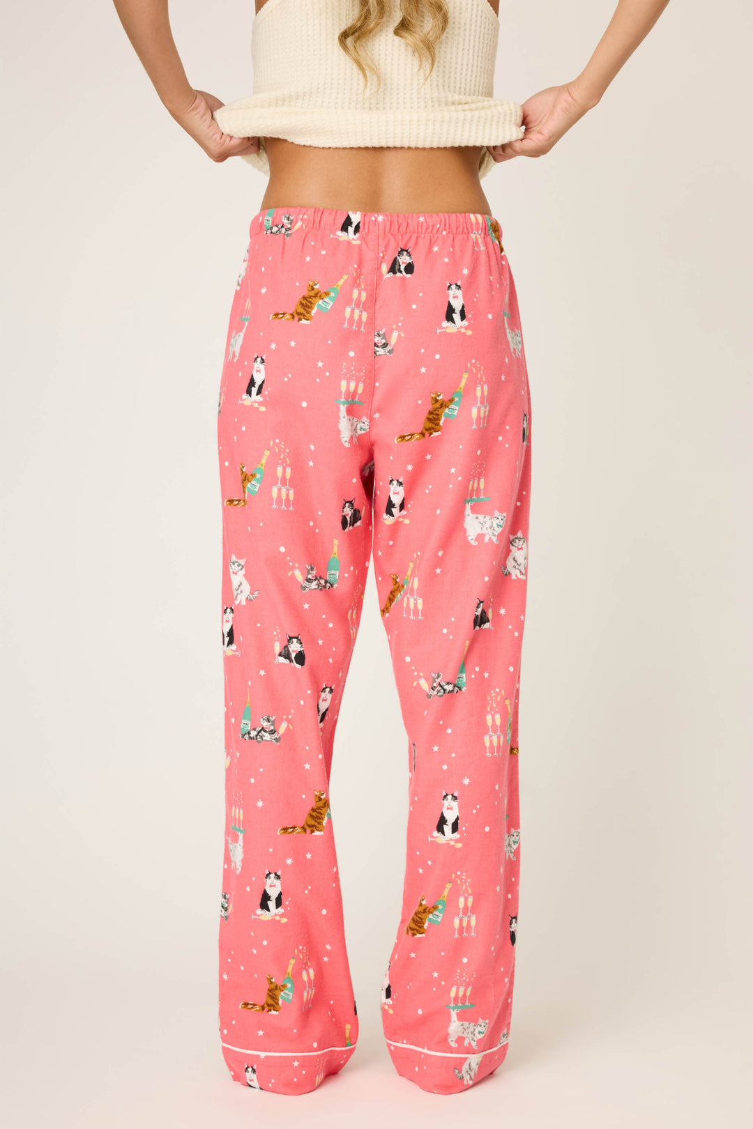 A person wears P.J. Salvage's Let's Get Fizzical Cat Print Flannel Pant, facing away and lifting their beige ribbed top to reveal the elastic waistband. The background is plain and light-colored.