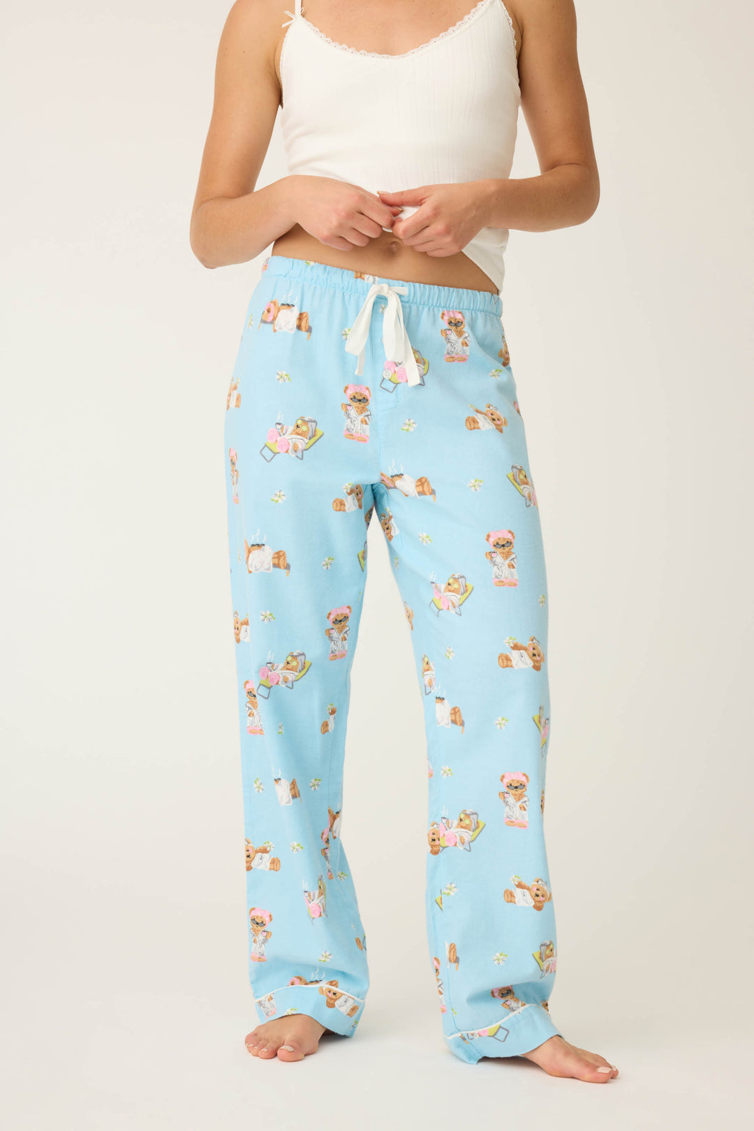 A person stands barefoot against a plain background wearing P.J. Salvage Beary Cute Bear Print Flannel Pants in light blue with a teddy bear print and a white tank top; their face is not visible.
