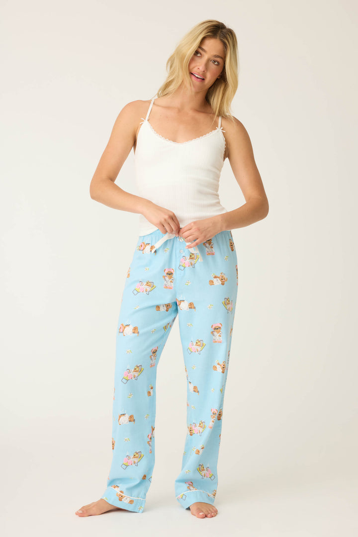 A woman stands barefoot against a plain background, wearing a white tank top and P.J. Salvage Beary Cute Bear Print Flannel Pants in light blue. She holds the waistband with both hands and gazes slightly to the side.