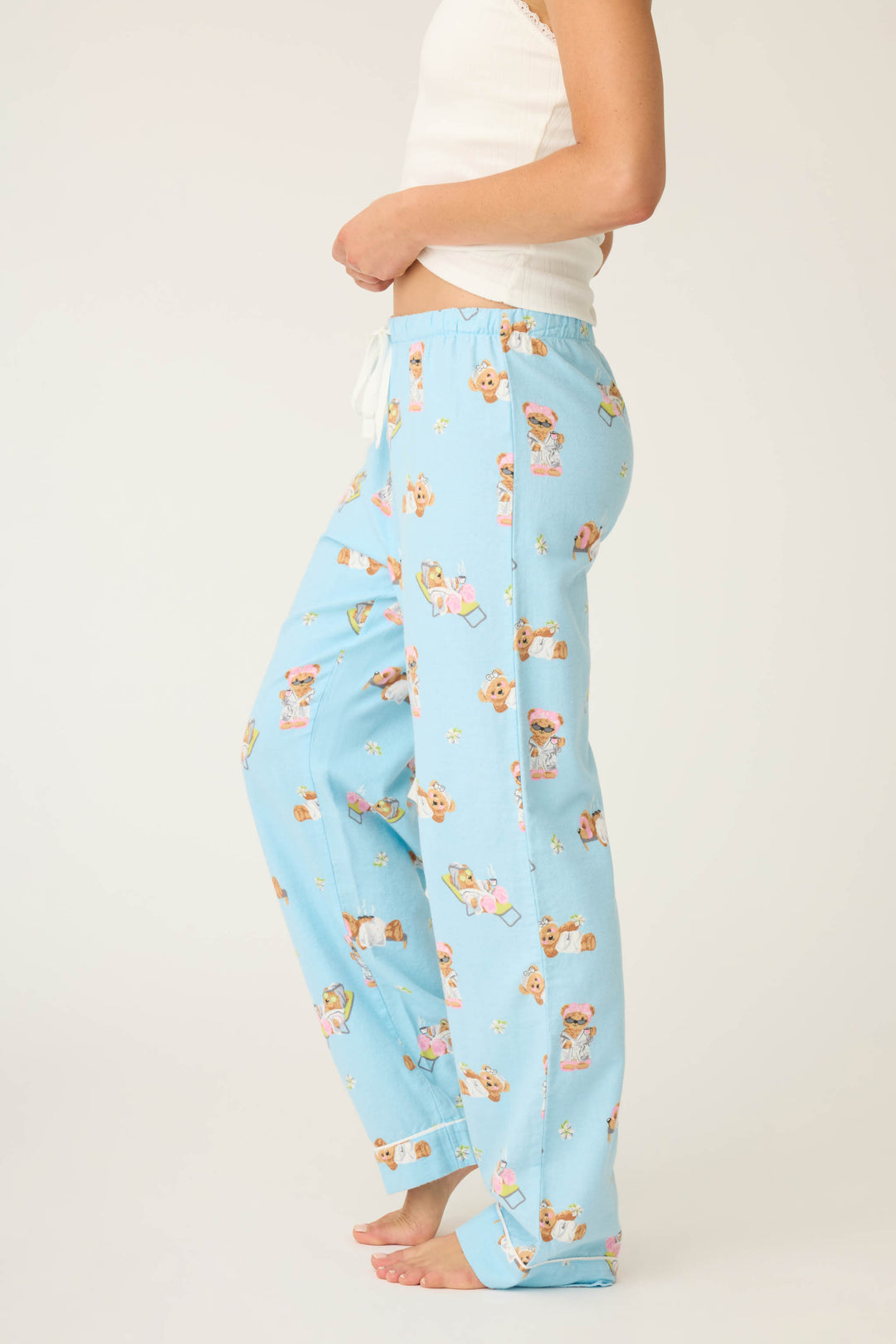 A person stands sideways barefoot against a plain, light background, wearing P.J. Salvage Beary Cute Bear Print Flannel Pants in light blue with teddy bears and books, paired with a white sleeveless top.