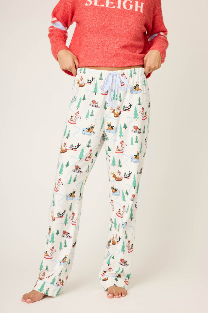 A person wears P.J. Salvage Sleigh All Day Winter Print Flannel Pants with festive animal and tree designs, paired with a red "SLEIGH" sweater, standing barefoot against a plain background.