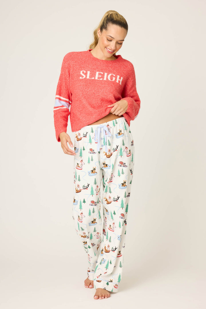 A smiling woman stands barefoot on a light background, wearing P.J. Salvage’s Sleigh All Day Winter Print Flannel Pant and a red “SLEIGH” sweater, holding the hem as she looks down.