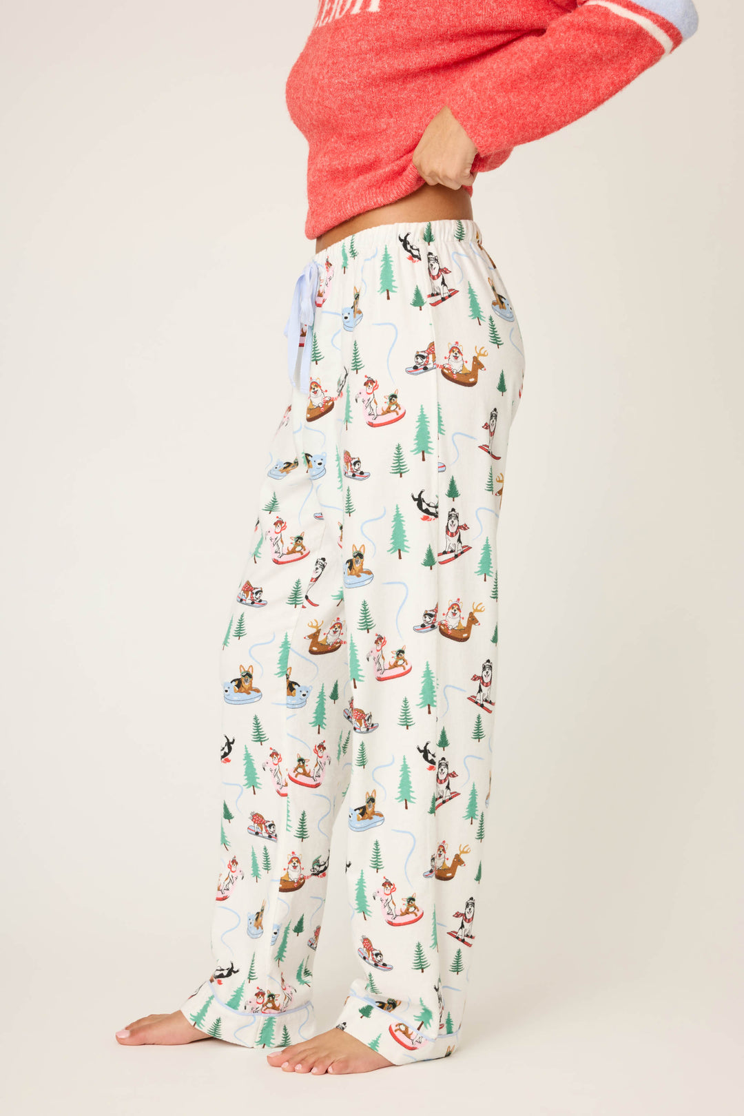 A person stands barefoot wearing P.J. Salvage Sleigh All Day Winter Print Flannel Pants and a red long-sleeve top. Their head is out of frame.