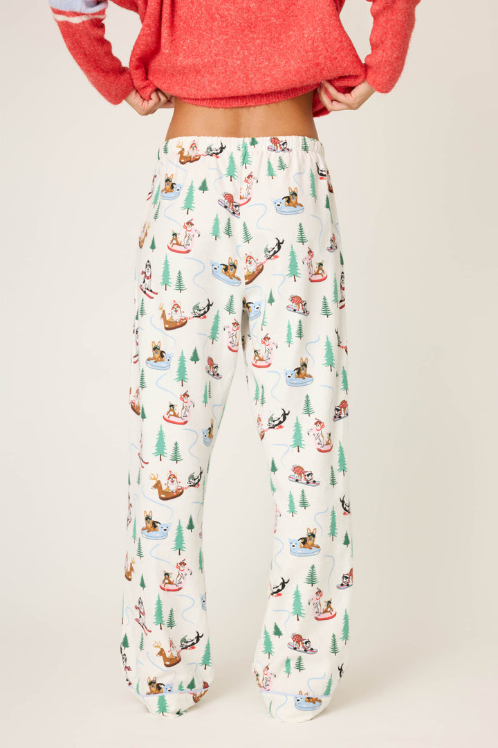A person models the P.J. Salvage Sleigh All Day Winter Print Flannel Pant, featuring a playful sledding dog and tree pattern, shown waist down against a plain background.