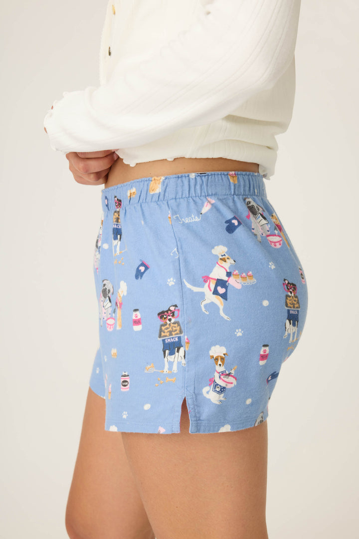 A person stands sideways wearing P.J. Salvage DOGS SHORT FLANNELS—light blue pajama shorts with cartoon dog and drink prints, small side slit, and elastic waistband—paired with a white long-sleeve top.